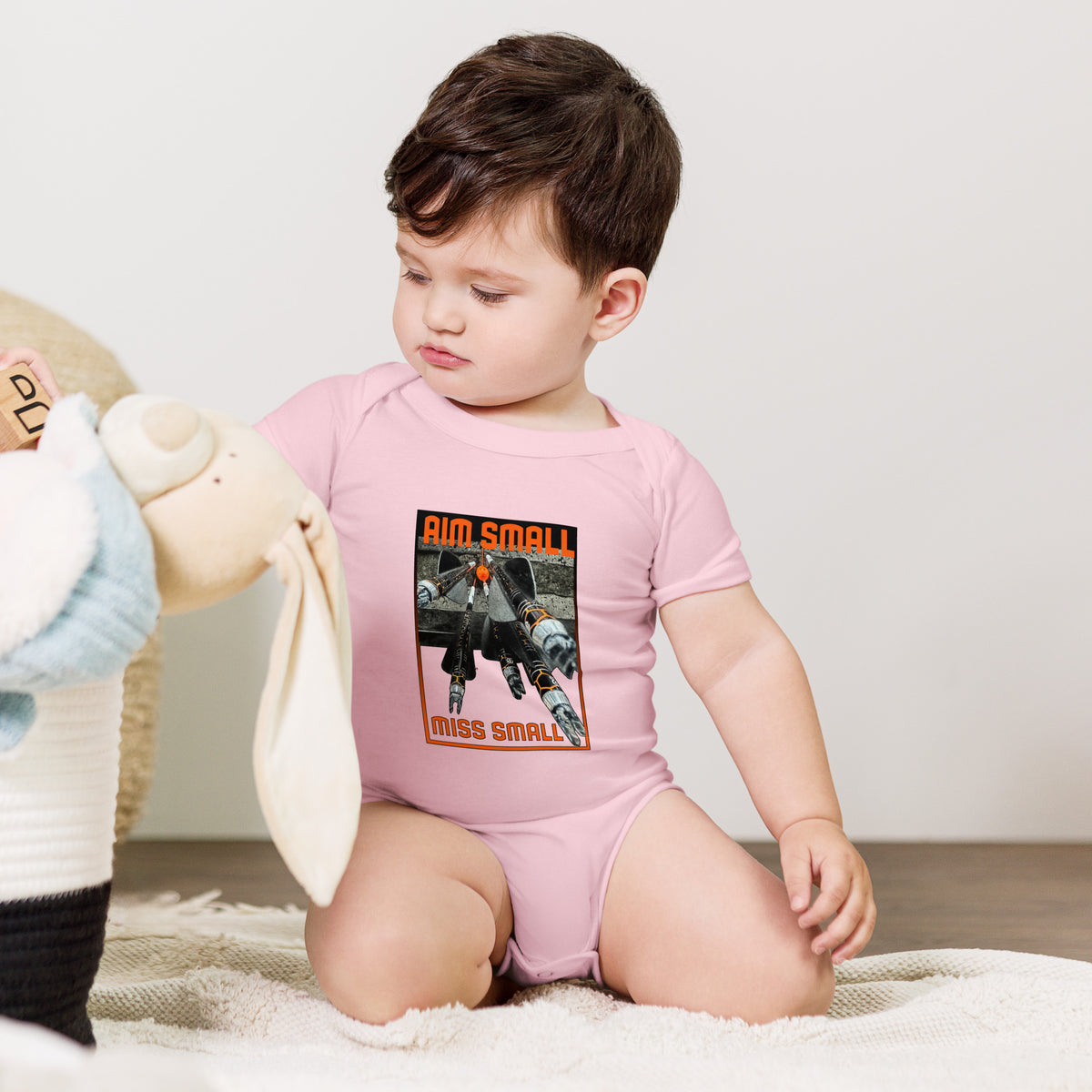 Aim Small Miss Small Baby Onesie in Pink, worn by a baby sitting with a plush toy
